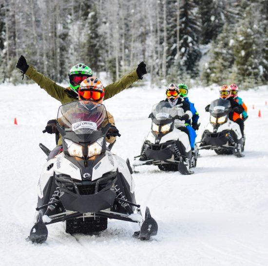 Three snowmobiles with two riders on each machine raising their arms in enjoyment as they navigate the practice course at Toby Creek Adventures
