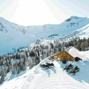 Ariel alpine view of the cozy Paradise Alpine Cabin with snowmobiles out front, smoke from the chimney at Toby Creek Adventures